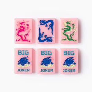 Puede incluir: Seis fichas rectangulares de color rosa dispuestas en dos filas. La fila superior presenta tres fichas con ilustraciones de dragones en rosa, azul y verde. La fila inferior presenta tres fichas id&eacute;nticas con un gr&aacute;fico de rana azul y el texto BIG JOKER en azul.
