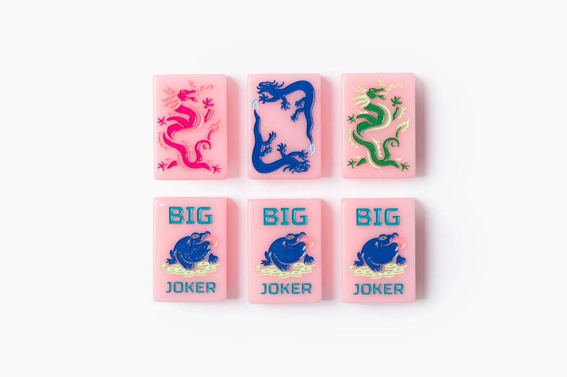 Puede incluir: Seis fichas rectangulares de color rosa dispuestas en dos filas. La fila superior presenta tres fichas con ilustraciones de dragones en rosa, azul y verde. La fila inferior presenta tres fichas id&eacute;nticas con un gr&aacute;fico de rana azul y el texto BIG JOKER en azul.