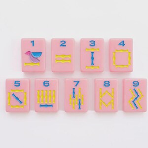 May include: A set of nine rectangular pink tiles arranged in two rows, each featuring a blue numeral from one to nine at the top. The tiles display various engraved patterns in yellow and blue, including geometric shapes, lines, and a bird icon.