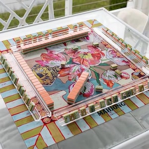 May include: A square mahjong game set featuring a colorful floral patterned mat with pink, red, and green tulips. The set includes clear acrylic tile racks and numerous mahjong tiles with peach-colored backs and green-patterned faces arranged on the mat.