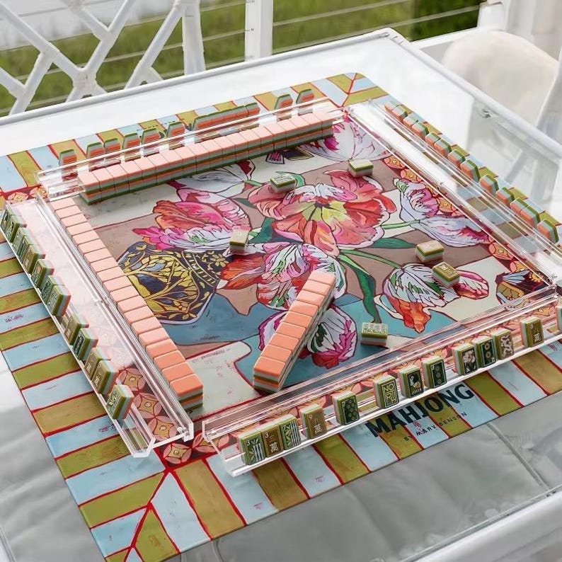 May include: A square mahjong game set featuring a colorful floral patterned mat with pink, red, and green tulips. The set includes clear acrylic tile racks and numerous mahjong tiles with peach-colored backs and green-patterned faces arranged on the mat.