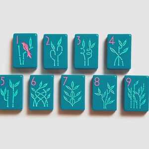 K&ouml;nnte beinhalten: Neun rechteckige, blaugr&uuml;ne Mahjong-Spielsteine in zwei Reihen. Jeder Stein zeigt eine pinkfarbene Ziffer von eins bis neun in der oberen linken Ecke und eine hellgr&uuml;ne Bambus-Illustration. Der erste Stein enth&auml;lt einen pinkfarbenen Vogel.