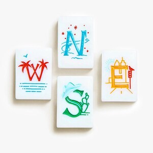May include: Four rectangular white tiles representing cardinal directions. The W tile features red palm trees, the N tile has a blue letter with red stars, the E tile displays a yellow city skyline with a bird, and the S tile shows a green letter with a cactus.