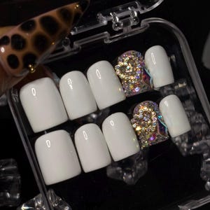 Puede incluir: Un juego de diez uñas postizas en un estuche de plástico transparente. Ocho uñas tienen un acabado blanco brillante sólido y forma cuadrada. Dos uñas están adornadas con un denso grupo de pedrería iridiscente y detalles de corazones dorados.
