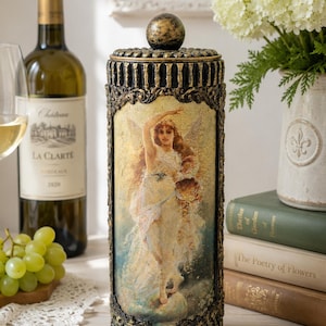 May include: A cylindrical decorative container with a black and gold finish, featuring a central image of a winged figure in a flowing white dress. The lid is topped with a spherical knob, and the exterior has ornate, textured vertical detailing.