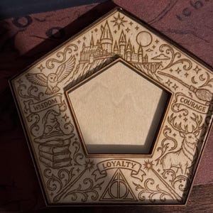 May include: A pentagonal wooden frame featuring laser-engraved illustrations including a castle, an owl, a sorting hat on books, a broomstick, and a stag. The words WISDOM, COURAGE, and LOYALTY are engraved on banners, along with a triangular symbol.