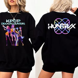 May include: Black sweatshirt with the text "KPOP DEMON HUNTERS" and a graphic of three anime-style figures. The back of the sweatshirt features the word "HUNTRX" with a colorful geometric design.
