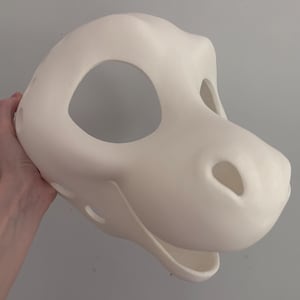May include: A blank, off-white dinosaur mask with large eye openings, a snout, and a wide open mouth. The mask is held by a hand and is made of a smooth, solid material. The mask is likely for costume or cosplay purposes.