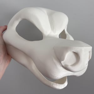 May include: A white, 3D-printed animal mask, featuring large eye openings, a prominent snout, and a detailed texture. The mask is held by a hand, showcasing its size and form. The design suggests a stylized animal head.