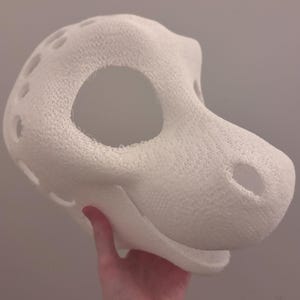 May include: A white, textured, 3D-printed mask in the shape of a stylized animal head. The mask has large eye openings, nostril holes, and several ventilation holes along the sides. The mask is held by a hand.