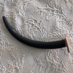 May include: A curved black furniture leg with a smooth finish, featuring a rectangular beige mounting plate at one end. The leg has a tapered, slender design and is positioned horizontally against a white textured fabric background.