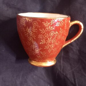 May include: A ceramic teacup featuring a deep red exterior with a repeating gold floral pattern. The cup has a gold-trimmed rim, a gold-painted handle, and a gold-finished circular base. The interior of the teacup is a solid white colour.