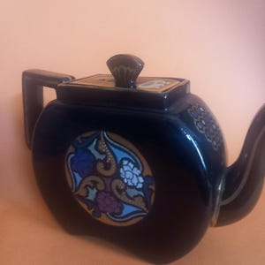 Peut inclure: Une théière en céramique bleu foncé avec un couvercle rectangulaire et une anse sur le côté gauche. Le corps présente un médaillon circulaire avec des motifs floraux complexes dans des tons de bleu, violet et or. Le bec verseur est courbé.
