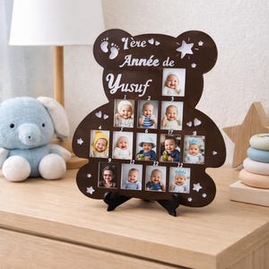 Peut inclure: Cadre photo en bois marron en forme d'ours, contenant douze petites photos d'un bébé. Le cadre porte l'inscription "1ère Année de Yusuf" en écriture blanche. Le cadre est posé sur une surface en bois.