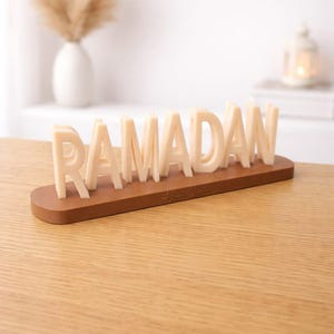 Peut inclure: Décoration de Ramadan avec le mot "RAMADAN" en lettres majuscules crème, montées sur une base ovale marron. L'ensemble est posé sur une surface en bois brun clair.
