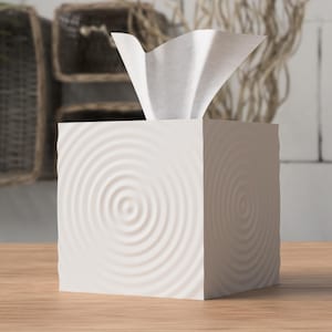 Peut inclure: Boîte à mouchoirs blanche avec un motif de cercles concentriques sur chaque face. La boîte carrée est posée sur une surface en bois. Des mouchoirs blancs dépassent du dessus. L'arrière-plan comprend des paniers tressés et des éléments décoratifs.