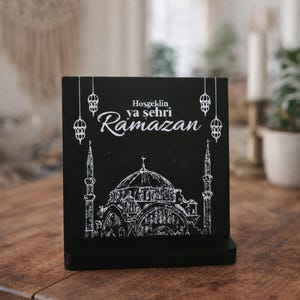 Könnte beinhalten: Schwarzes Holzschild mit einer weißen Moscheedarstellung und dem Text "Hosgeldin ya şehri Ramazan". Das Schild hat hängende Laternen-Designs. Es wird auf einem Holztisch präsentiert.