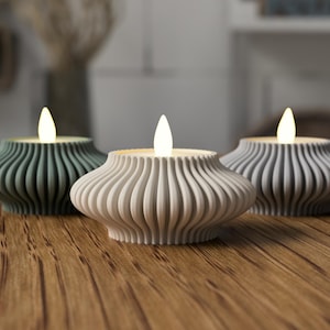 Peut inclure: Trois bougies LED décoratives avec une lueur chaleureuse. Les bougeoirs sont dans des tons de vert, blanc et gris, avec un design arrondi et nervuré. Elles sont placées sur une surface en bois.