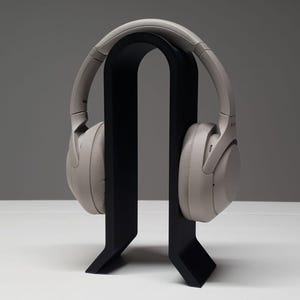 Peut inclure: Casque audio gris clair posé sur un support noir. Le support, de conception minimaliste, présente un sommet incurvé et une base robuste, offrant une présentation élégante pour le casque.
