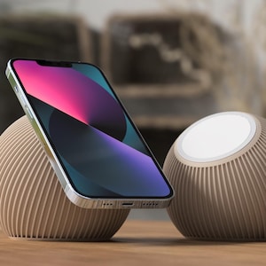 Könnte beinhalten: Ein silbernes Smartphone mit farbenfrohem Bildschirm liegt auf einem beigefarbenen, gerippten, kugelförmigen Ständer. Rechts befindet sich ein weiterer ähnlicher Ständer mit weißer kreisförmiger Oberseite. Die Gegenstände befinden sich auf einer Holzoberfläche.