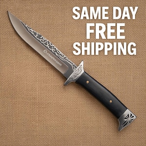 May include: A large, silver-colored knife with a decorative etched blade and a black handle. The knife features a guard with an ornate design and a decorative end cap. The text "Garantom" is etched on the blade.