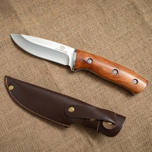May include: A stainless steel hunting knife with a wooden handle and a brown leather sheath. The knife blade is silver and the handle is a warm brown color. The sheath is a darker brown with a snap closure.