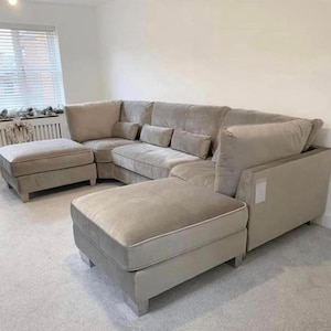 May include: A large, beige sectional sofa with a chaise lounge and several cushions. The sofa is upholstered in a soft, velvet-like fabric. It sits on a light grey carpet. The sofa is designed for comfort.