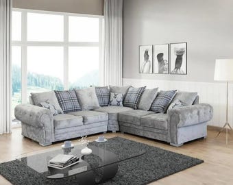 May include: A large, grey corner sofa with patterned cushions, set in a living room. The cushions feature plaid and floral designs. A dark grey rug and glass coffee table are in front of the sofa. The room has a large window and artwork.