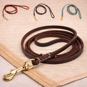 May include: A collection of leather dog leashes in various colors: dark brown, red, black, and teal. Each leash features a brass-colored clasp. The leashes are displayed on a neutral-toned fabric background.