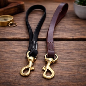 May include: Two leather wrist straps with brass-colored clasps. One strap is black, the other is brown. The straps are looped and stitched, designed for attaching to a bag or other item. The background is a wooden surface.
