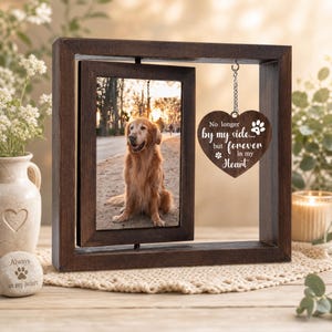 Op de afbeelding: Een donkerbruine houten draaibare fotolijst met een foto van een gouden hond. Een hartvormig houten ornament hangt aan een zilveren ketting aan de bovenkant van de lijst, met de witte tekst: No longer by my side... but forever in my heart.