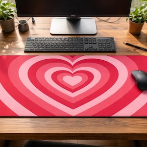 Peut inclure: Tapis de bureau rouge et rose en forme de cœur. Le motif présente des cercles concentriques de différentes nuances de rouge, formant un cœur. Une souris d'ordinateur noire est posée sur le tapis, avec un clavier et un écran en arrière-plan.