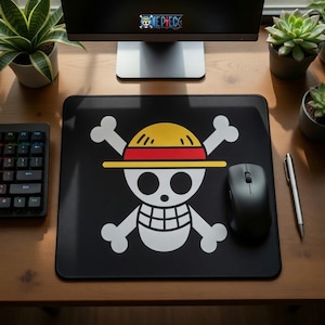 Puede incluir: Alfombrilla de ratón rectangular negra con el icónico logotipo de calavera y huesos cruzados de la serie de anime One Piece. El logotipo es blanco, amarillo y rojo. Un ratón de ordenador negro y un bolígrafo plateado están sobre el escritorio de madera.