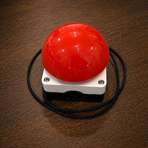 May include: A large, glossy red dome-shaped button mounted on a square white and black base. The device features a thin black circular cord that loops around the base. The construction includes visible metal screws at the corners of the white top.