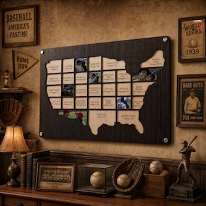 May include: A dark wood wall display shaped like the United States map featuring a grid of square slots for photos of baseball stadiums. The board is labeled MLB STADIUM TOUR and includes text for various stadium names and locations across the map.