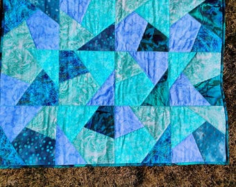 May include: A handmade quilt featuring a geometric pattern of triangles and squares in shades of blue and teal. The quilt has a teal border and is made of various patterned fabrics, creating a textured appearance.