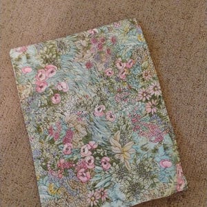 May include: A rectangular fabric item with a floral pattern. The design features pink roses, white and yellow flowers, and green foliage on a light blue background. The item is set against a neutral brown surface.