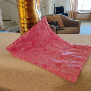 May include: A pink decorative cloth with a white floral and swirl pattern. The cloth is folded and placed on a beige surface. A tall, gold-coloured vase is in the background.