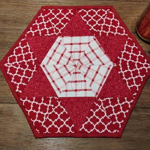 Può includere: Un sottopentola esagonale trapuntato rosso e bianco. Il sottopentola presenta un esagono centrale con un motivo a quadri bianchi e rossi e ricami floreali. Le sezioni esterne hanno uno sfondo rosso con disegni a quadrifoglio bianchi.