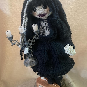 May include: A handmade crochet doll with black hair and a black dress, holding a candelabra and a bouquet of white roses. The doll has a pale face, fangs, and is standing on a small wooden coffin. The doll is approximately 25 cm tall.