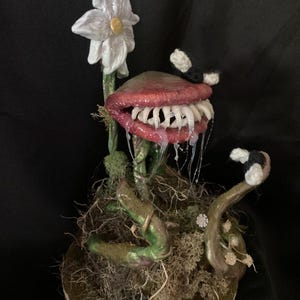 Op de afbeelding: Een handgemaakt keramisch beeld van een vleesetende plant met een grote, open mond, scherpe tanden en een druipende rode tong. De plant heeft een witte bloem en groene ranken en staat op een basis van mos en takken.