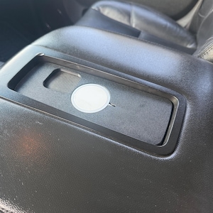 Puede incluir: Consola de coche de cuero negro con una bandeja incorporada. La bandeja tiene una pequeña pegatina redonda blanca y una ranura para una herramienta. La consola forma parte del interior de un coche.