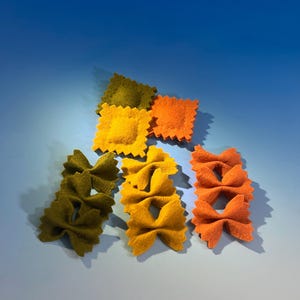 May include: An assortment of colorful felt pasta shapes, including ravioli and farfalle, in shades of green, yellow, and orange. The pasta is arranged on a gradient blue background, creating a playful and artistic display.