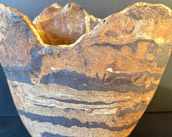 Pode incluir: Uma tigela de cerâmica moldada à mão com uma borda irregular e ondulada. O exterior apresenta uma superfície texturizada e terrosa com faixas horizontais e giratórias de castanho escuro, bege e creme, criando um aspeto orgânico.