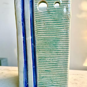 May include: Rectangular ceramic vase with a textured surface. The vase features vertical blue stripes on one side and horizontal green stripes on the other. Two circular holes are near the top.