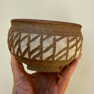 May include: A round, footed ceramic bowl with a textured, dark brown speckled clay body. The exterior features a wide band of light gray, matte-finished clay with a repeating, incised geometric pattern of diagonal lines forming diamond shapes.