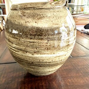 May include: A large, round, ceramic pottery vessel with a lid. The pottery is a mottled mix of tan, brown, and cream colors. The vessel has a wide opening and a handle on the side of the lid. The pottery sits on a dark wooden surface.
