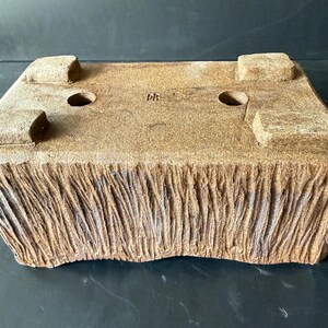 Pu&ograve; includere: Un vaso rettangolare in ceramica marrone per bonsai con una finitura esterna ruvida che ricorda la corteccia. La base presenta quattro piedini robusti, due fori di drenaggio circolari e le lettere DB stampate al centro.