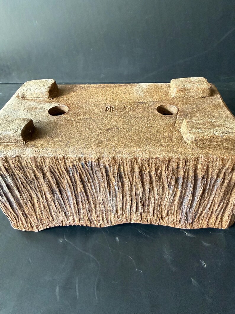 Pu&ograve; includere: Un vaso rettangolare in ceramica marrone per bonsai con una finitura esterna ruvida che ricorda la corteccia. La base presenta quattro piedini robusti, due fori di drenaggio circolari e le lettere DB stampate al centro.