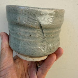 May include: A cylindrical ceramic cup with a light sage green glaze and a speckled finish. The exterior features horizontal ridges and a prominent diagonal textured pattern of incised lines. The base is unglazed, revealing a light beige clay body.